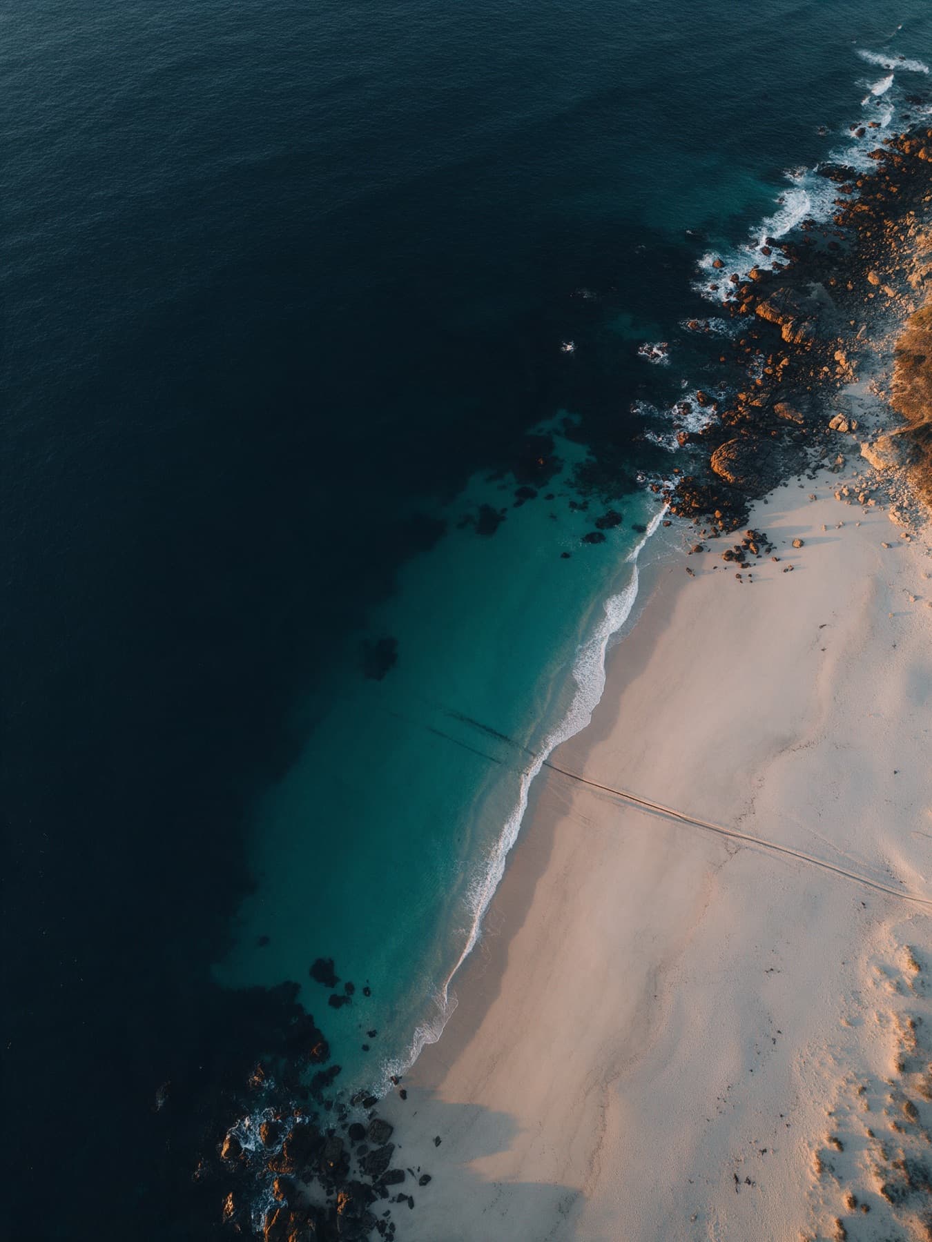 Aerial view of a coastline where turquoise shallows meet deep water, with a faint cable trench crossing the beach